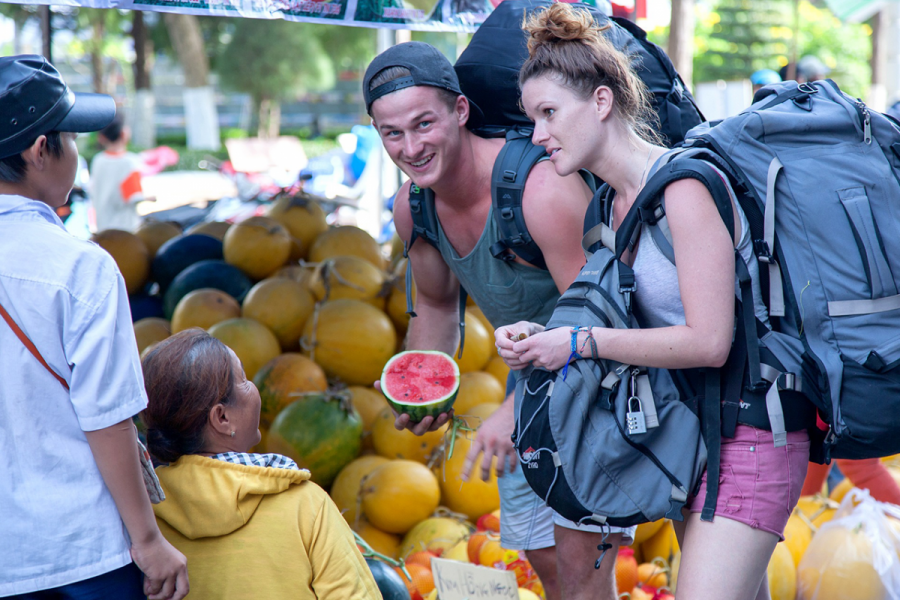 Two people with backpags holding fruit, speaking to a market stall operator