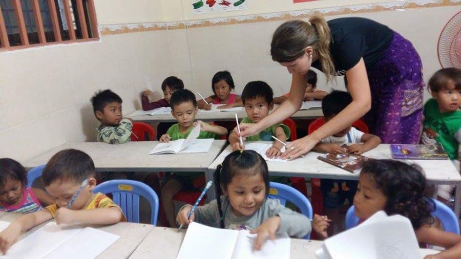 Kids sitting at desks in a classroom with a teacher helping a student