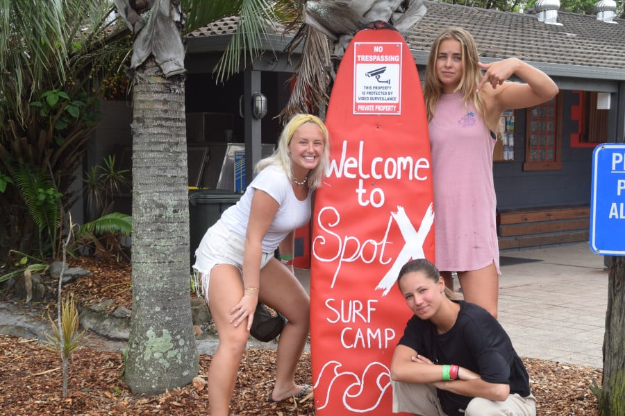A group of travellers pose next to a sign shaped like a surfboard that reads 'Welcome to Spot X Surf Camp'